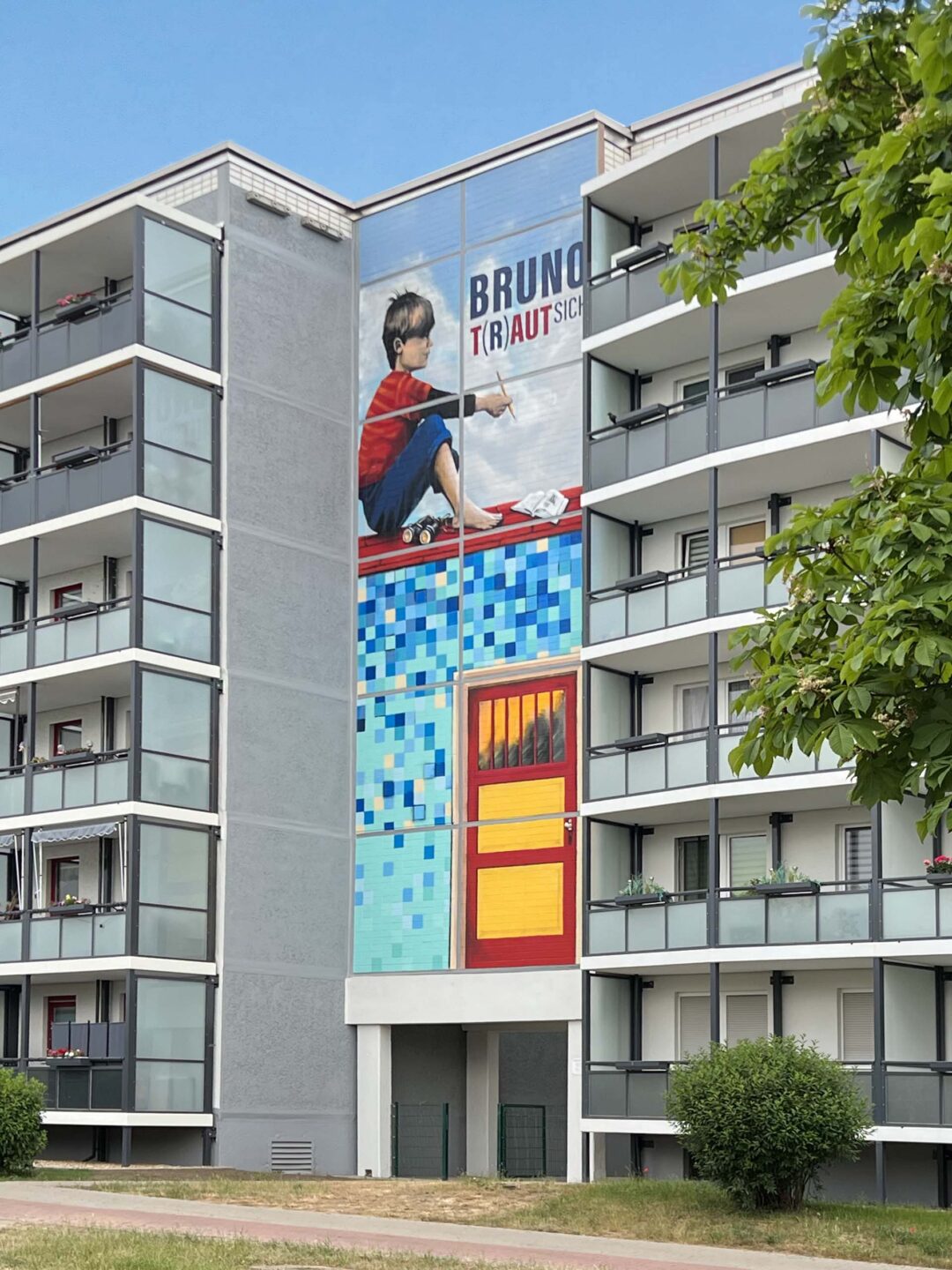 Hausfassade mit vielen Balkonen in der Mitte befindet sich ein großes Graffiti mit dem Motiv eines Jungen der auf einem Häuserdach sitzt und nachdenkend in die Ferne schaut