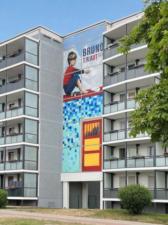 Hausfassade mit vielen Balkonen in der Mitte befindet sich ein großes Graffiti mit dem Motiv eines Jungen der auf einem Häuserdach sitzt und nachdenkend in die Ferne schaut